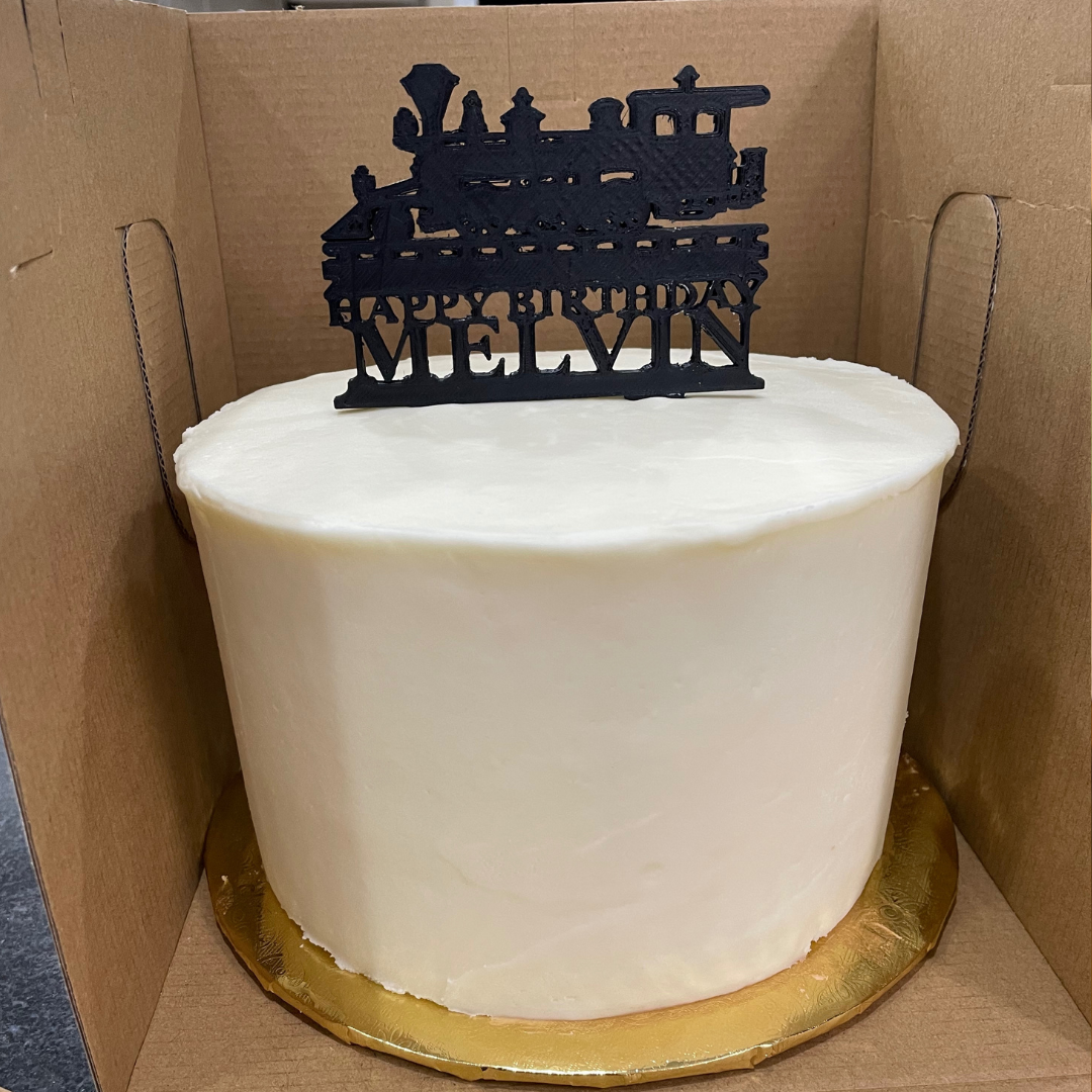 White frosted Carrot Cake with a black train silhouette topper reading “Happy Birthday Melvin.”
