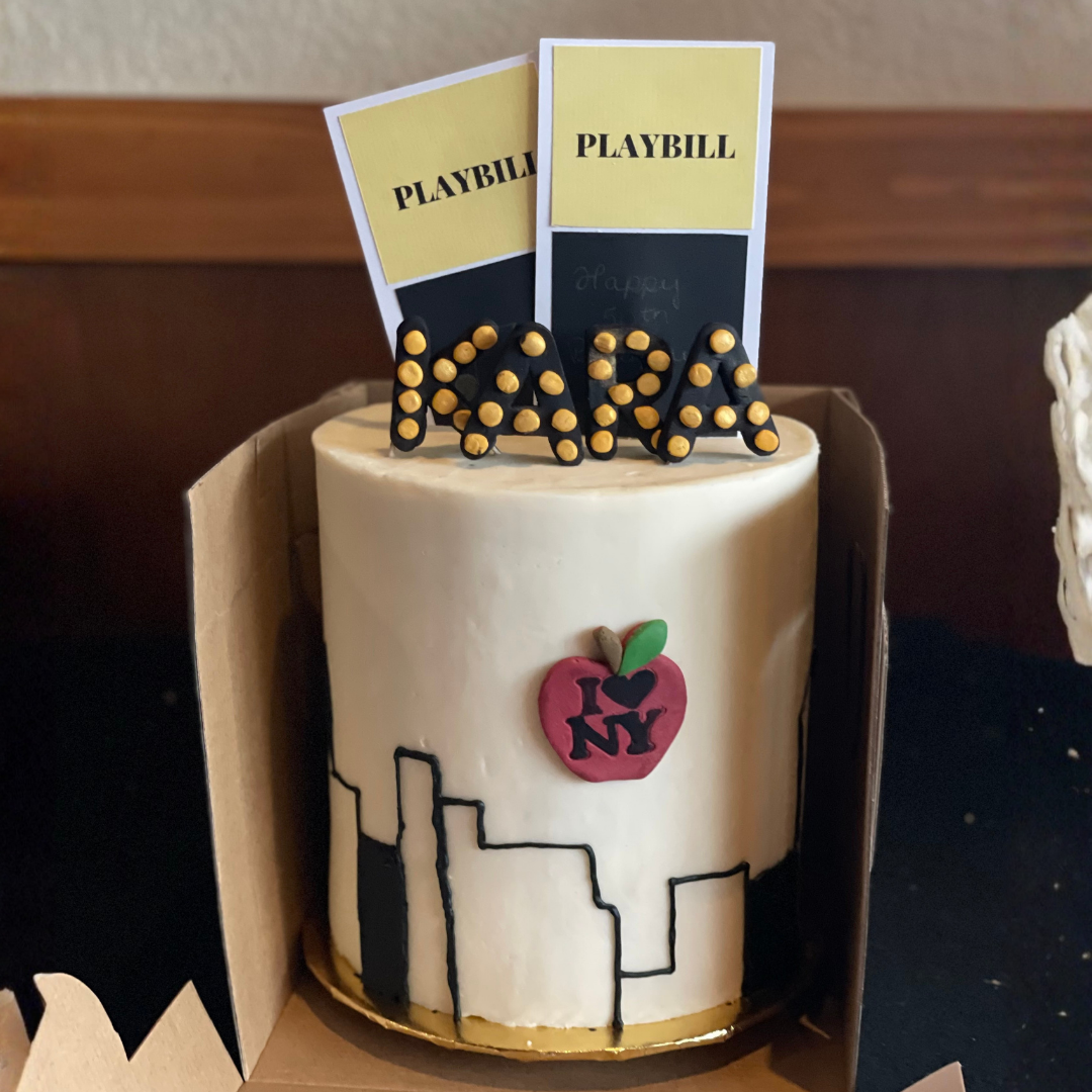 White cake decorated with a black skyline, red apple, and yellow “Playbill” toppers.