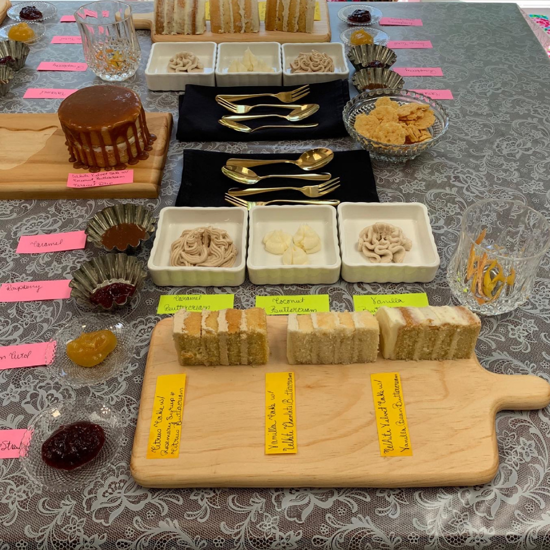 A wedding cake tasting setup featuring slices of layered cake, buttercreams, and fillings arranged on wooden boards and labeled with colorful tags.