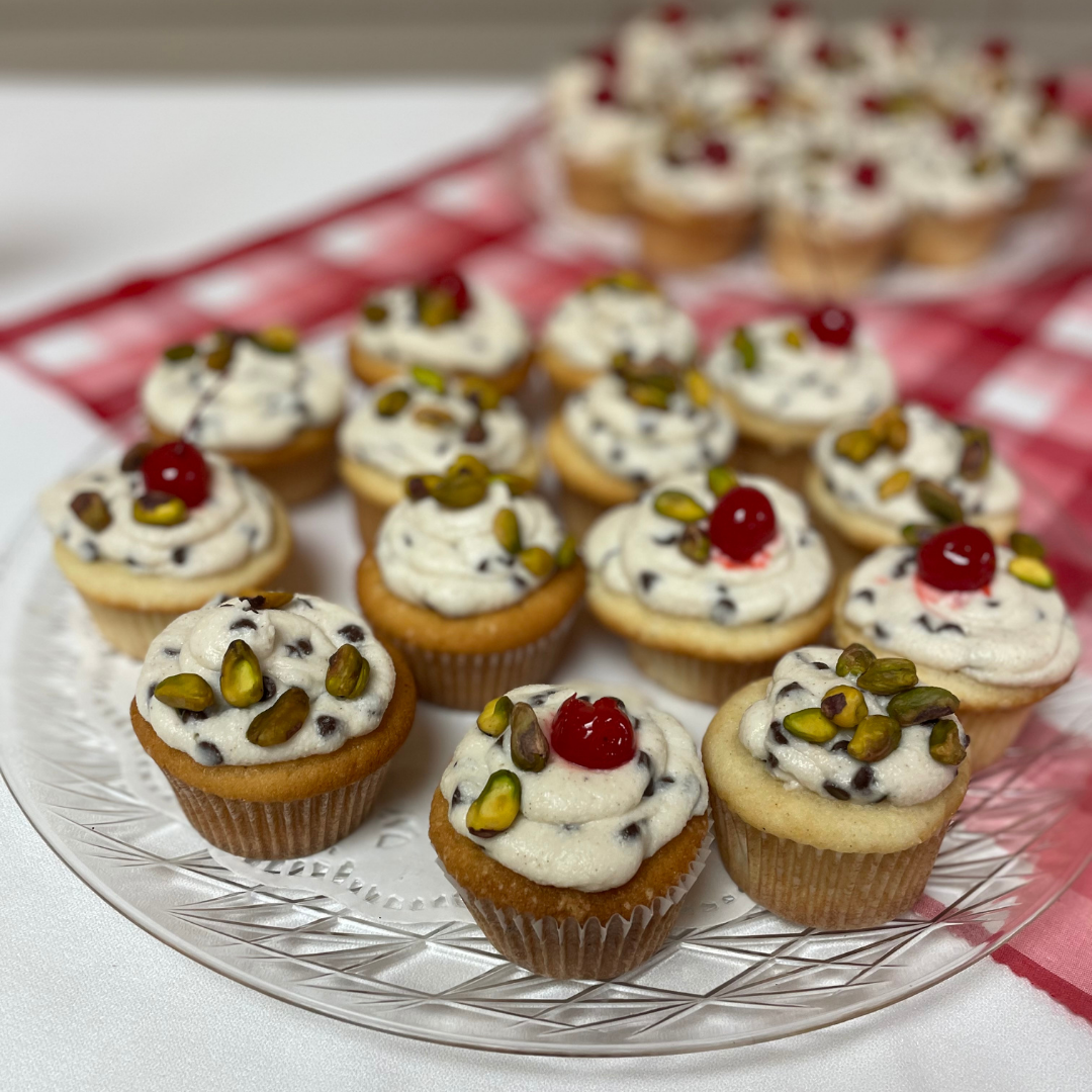 anilla cupcakes topped with chocolate chip cannoli cream, crushed pistachios, and a maraschino cherry.