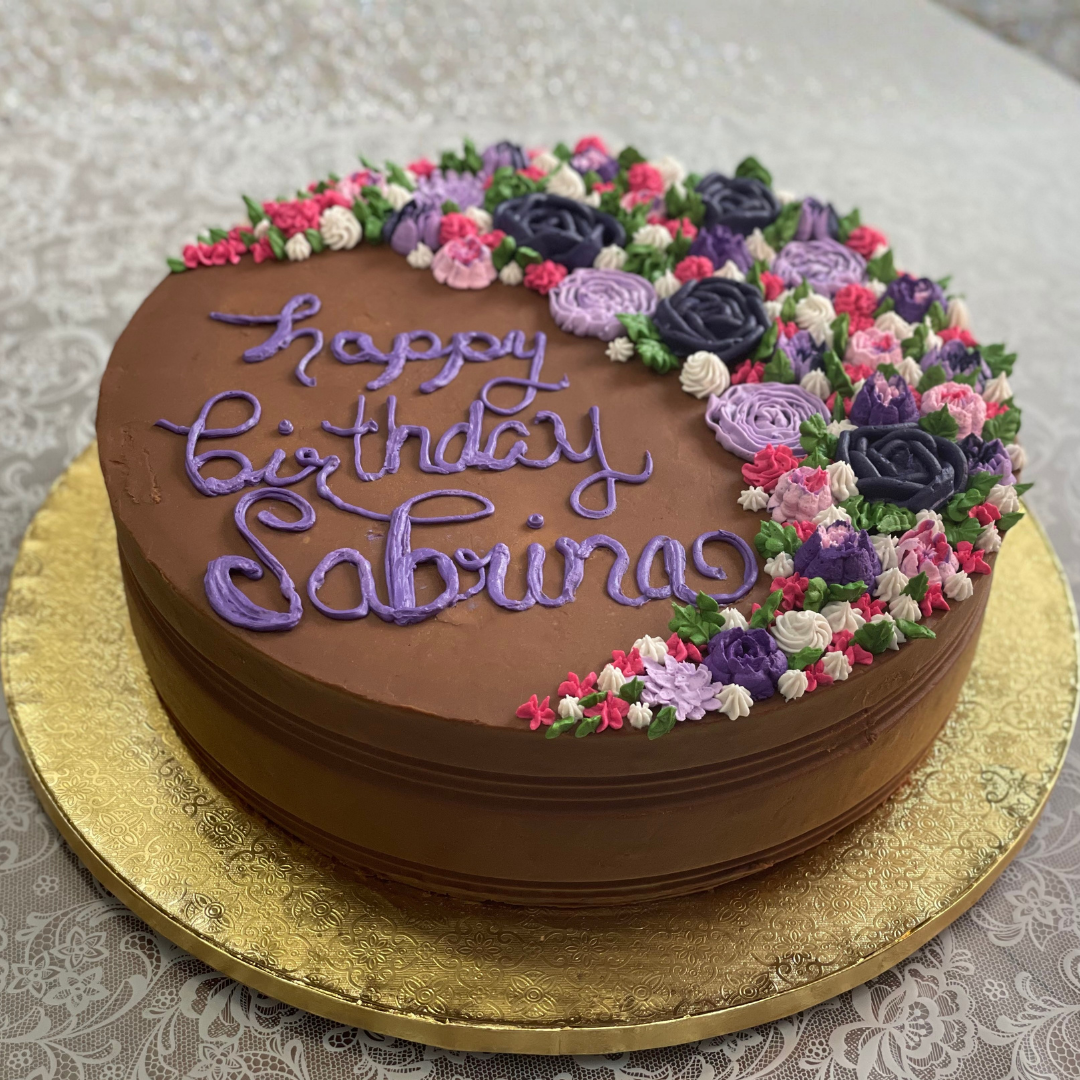 Chocolate birthday cake with purple and pink floral buttercream decorations and “Happy Birthday Sabrina” written in lavender icing.