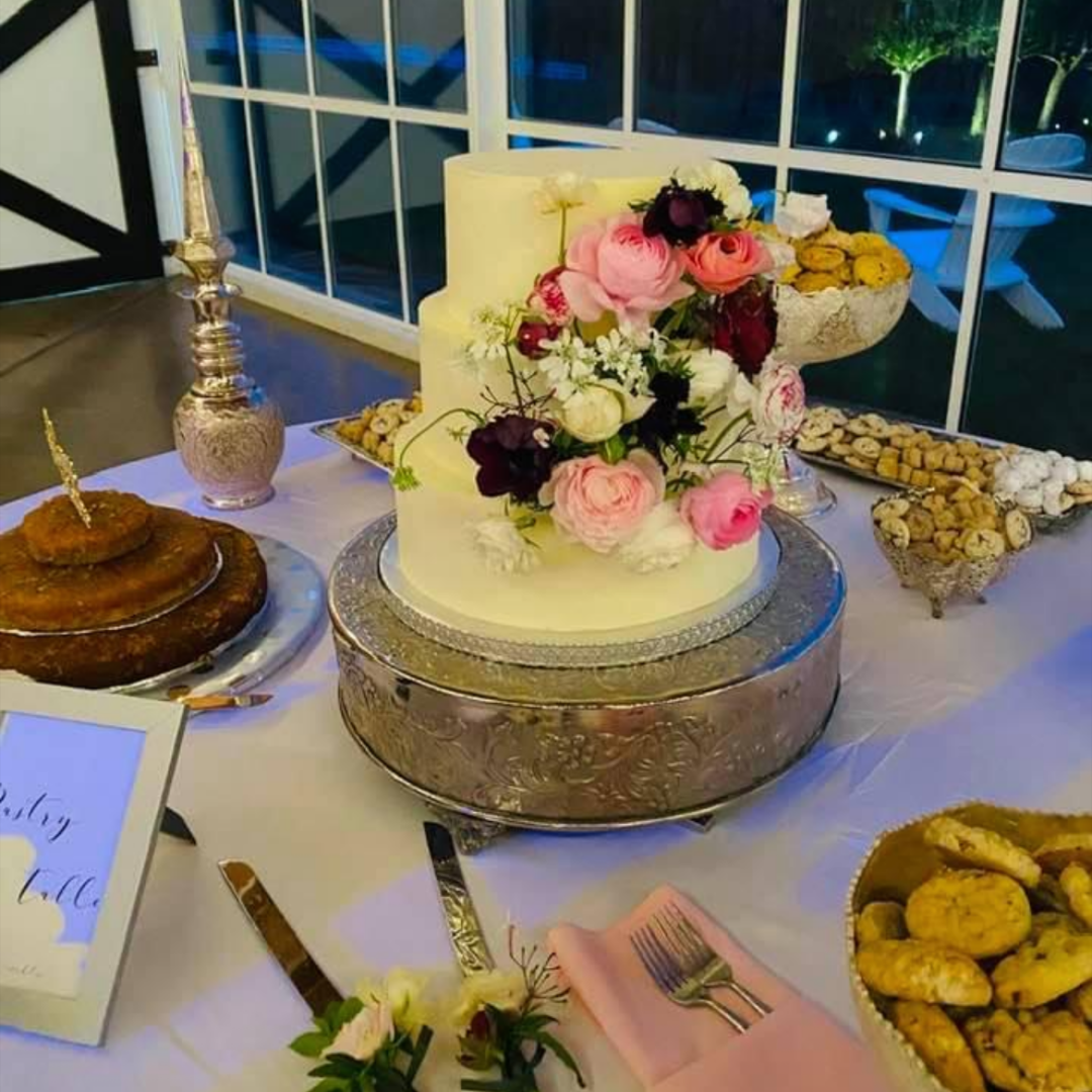 A three-tier white wedding cake with cascading floral arrangement surrounded by pastries on silver stands.