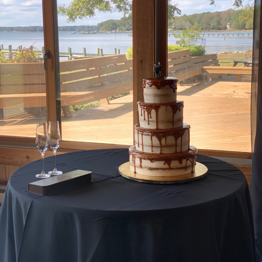 A four-tier naked-style cake with caramel drip icing set beside a lakeside window.