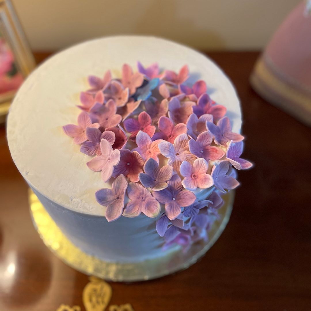 White buttercream cake topped with handmade sugar flowers in pastel pink and purple.