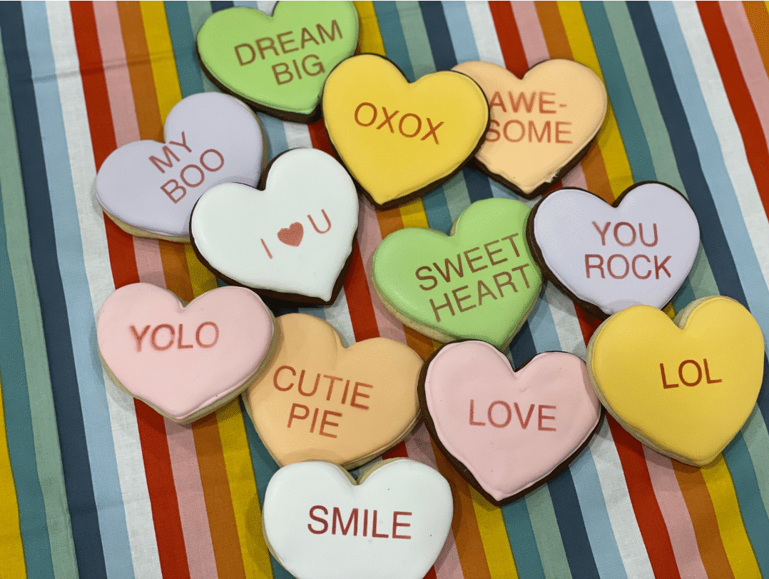 Heart-shaped royal icing cookies in soft pastel colors with classic and modern sayings like “Be Mine,” “You Rock,” “LOL,” “YOLO,” and “My Boo.”